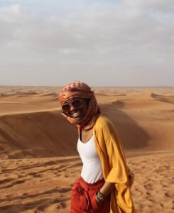 College student walks in front of a sand dune with a scarf wrapped around their head and hands in their pockets.