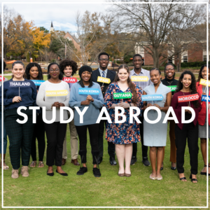 Image of several college students lined up in an outdoor setting facing the camera. Each student holds a sign with a couImage of three adults standing and looking at an old ledger in an archive room with shelves full of old documents. The image is a button link to the Undergraduate Research subpage.ntry name that is their study abroad destination. Image is a button linking to the Study Abroad subpage.